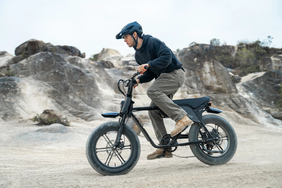 A helmeted rider on a TXED Maverick on a sandy track beside rocks.