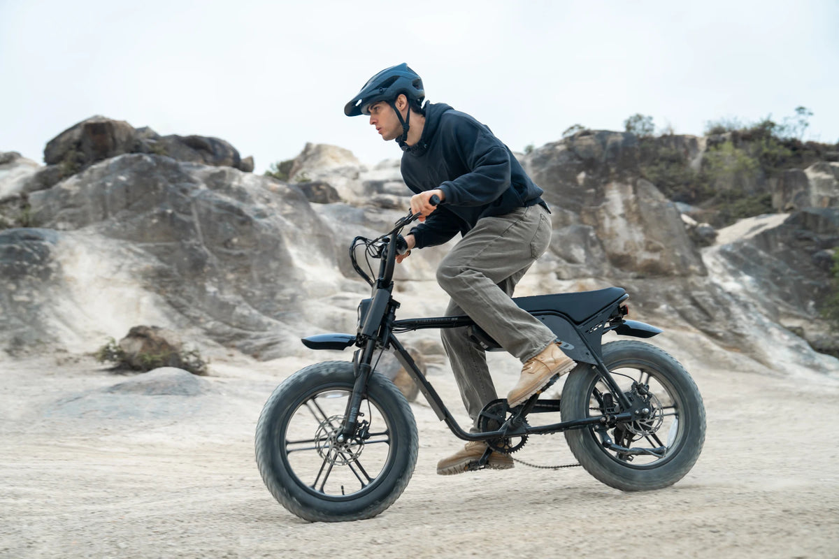 A helmeted rider on a TXED Maverick on a sandy track beside rocks.